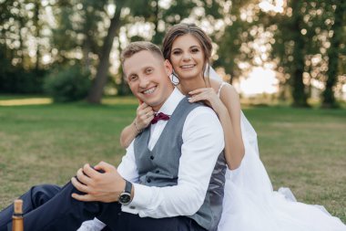 Yazın parkta yeşil çimlerde oturan güzel bir çiftin portresi. Genç, neşeli gelin arkadan genç ve yakışıklı damada sarılıyor. İlişki, aşk, kutlama..