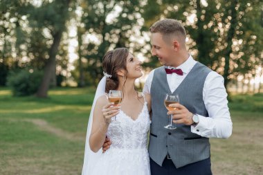 Mutlu evlilik çifti yazın parkta dikilirken yan görüntüsü. Genç bir damat, genç ve güzel bir kadına sarılıyor, elinde şarap kadehleri, gözleriyle buluşuyor. İlişki, aşk, kutlama..