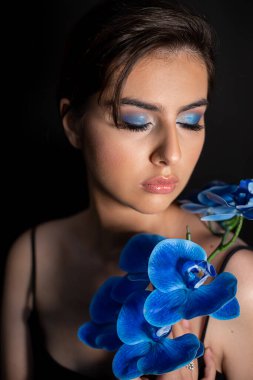 Dikey çekici, çekici, romantik esmer kadın mükemmel cilt ve mavi çiçek dalı ile makyajlı siyah arka planda aşağı bakıyor. Deri ve vücut bakımı. Kokulu kozmetik ürünü