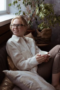 Dikey pozitiflik, gözlüklü, gülümseyen sarışın kadın. Çiçeğin yanındaki rahat koltukta oturuyor ve elinde kupa tutuyor. Evde kahve ya da çay içiyor. Rahatlamak ve fikir üretmek, sıcak atmosferde ilham vermek.