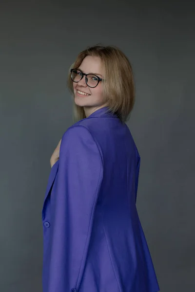 長い公正な髪を眼鏡をかけて若い笑顔の美しい女性のサイドビューでは 青いジャケットに投げ 手でエッジを保持し 灰色の背景に立っている スタジオ ファッション — ストック写真