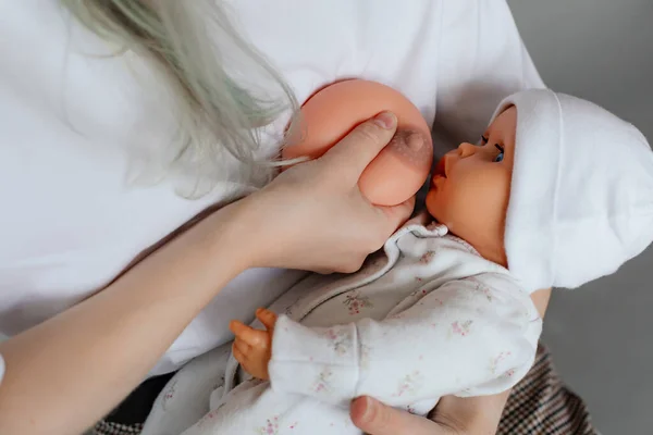 Cropped photo of young woman holding baby doll, showing how to feed baby with artificial fake breast silicone model on grey background. Childbirth, breastfeeding, breast model, fake, education.