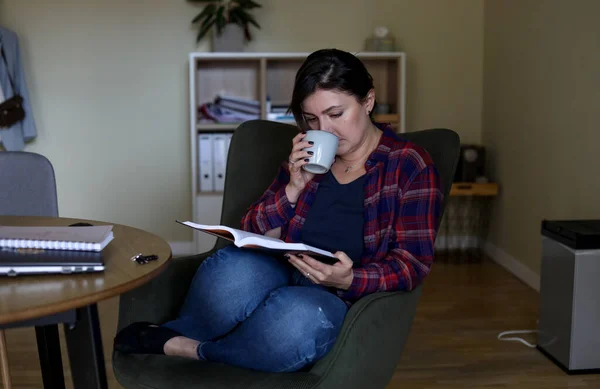 Calm, tired and pensive mature woman drinking tea and reading book at home. Education and study for adults. Relaxing and resting alone, time with yourself. Prepare to exam, adult courses for new job