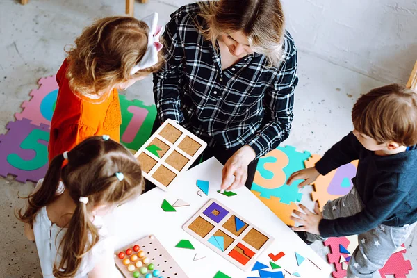 Development class for little children. Female teacher with children having fun and do smart puzzle with pieces exercises in kid development childcare center for preschoolers. High angle view
