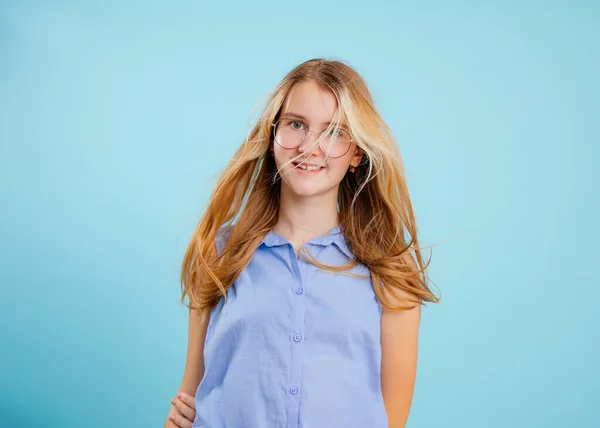 Smiling, positive teenage blonde girl in glasses in blue shirt with trendy hairstyle on blue isolated background. Bad vision, corrective eyeglasses, spectacles. Optical problems and check. Copy space