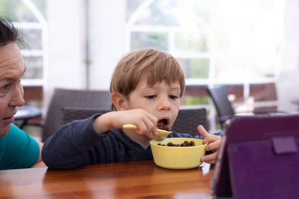 Little boy sit on chair at table and with interest watch cartoon on tablet computer with mother. Kid of kindergarten age open mouth to eat chocolate balls with milk. Ready breakfast, gadget addiction.