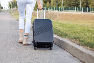 Slim legs of woman with suitcase walk along roadside of empty highway moving away while wait passing car. Lady escape from city to go anywhere. Travelling, free transportation, hitchhiking, vacations. clipart