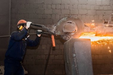 Koruyucu miğferli, solunum cihazlı bir erkek işçi, endüstriyel bir fabrikanın atölyesindeki uçan kıvılcımlarla dökme demir beton borular için ağır öğütme ekipmanlarını yönetiyor..