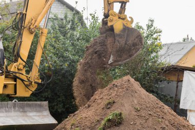 Bir kova kazıcı toprağı kırsal bir sanayi bölgesinin arka planına karşı bir yığın halinde döker. İnşaat alanında buldozer işi.