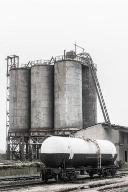 Demiryolu endüstriyel istasyonunun raylarında bulunan gübre tankı terk edilmiş bir fabrikadaki eski kum depolama tanklarının arka planına karşı..