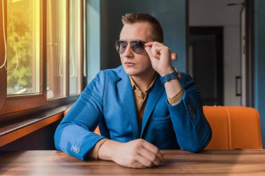 Kafkas görünümlü genç iş adamı şık bir portre, güneş gözlüğünü elleriyle ayarlıyor, pencereden dışarı bakıyor ve masada oturuyor..