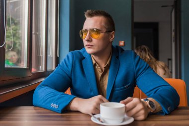 Düşünceli, genç, beyaz görünümlü, güneş gözlüklü ve takım elbiseli bir işadamı kahve molasında bir kafedeki bir masada oturur ve pencereden dışarı bakar..