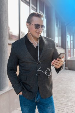 Ceket ve gömlek tarzında Avrupalı genç ve çekici bir işadamı güneş gözlüğü takıyor ve açık havada bir akıllı telefondan kulaklıkla müzik dinliyor..