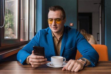 Avrupa görünümlü, güneş gözlüklü, ceketli ve gömlekli şık bir işadamı kahve molasında bir masada oturuyor, elinde bir akıllı telefon ya da cep telefonu tutuyor..