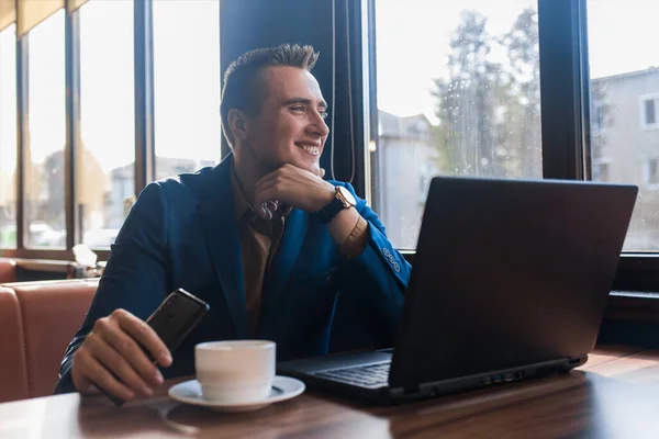 A business smiling man positive businessman a stylish of Caucasian appearance in a jacket, works in a laptop or computer, sitting at a table by the window in a cafe.