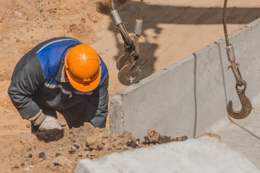 Tulumlu ve koruyucu başlıklı bir endüstri işçisi çelik bir halatla çelik bir kanca kaldırma mekanizmasını beton bir yapıya bağlar..