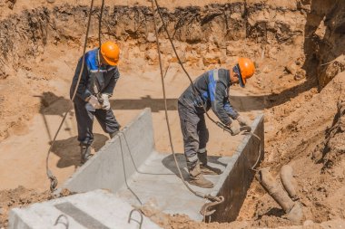 Belarus, Minsk bölgesi - 20 Mayıs 2020: Tulumlu ve koruyucu kasklı bir endüstri işçisi çelik bir halatla beton bir yapıya ağır demir kanca kaldırma mekanizması takıyor.