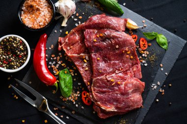 Baharatlı çiğ biftek ve siyah arka planda otlar. Tuz, biber, sarımsak, kırmızı biber ve biberiye.