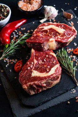 Baharatlı çiğ biftek ve siyah arka planda otlar. Tuz, biber, sarımsak, kırmızı biber ve biberiye.