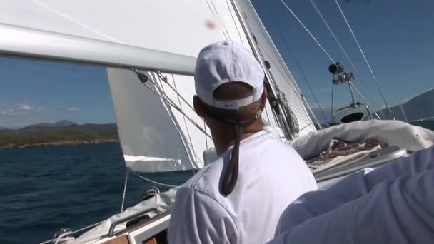 Человек в белой кепке и белой футболке поворачивает лебедку и веревки на яхте с белыми парусами. — стоковое видео