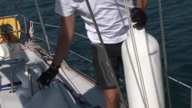 Beyaz şapkalı ve beyaz tişörtlü adam beyaz yelkenli yatta vinç ve ipleri çeviriyor..