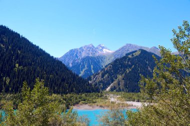 Manzaralı dağ gölü manzarası ve güneşli yaz gününde köknar ağaçlarıyla kaplı dağların yamaçları. Kazakistan 'daki Issyk Gölü' nün yatay fotoğrafı.