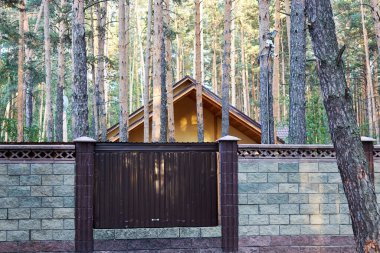 Çam ağaçlarının arasında beton blokların arkasında duran bir evin çatısı.