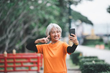 Yaşlı beyaz saçlı Asyalı kadın sabah parkta egzersiz yaparken görüntülü konuşuyor..