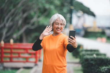 Yaşlı beyaz saçlı Asyalı kadın sabah parkta egzersiz yaparken görüntülü konuşuyor..