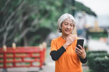 Yaşlı beyaz saçlı Asyalı kadın sabah parkta egzersiz yaparken görüntülü konuşuyor..