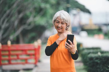 Yaşlı beyaz saçlı Asyalı kadın sabah parkta egzersiz yaparken görüntülü konuşuyor..