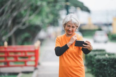 Yaşlı beyaz saçlı Asyalı kadın sabah parkta egzersiz yaparken görüntülü konuşuyor..