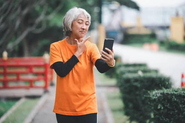 Elderly white haired Asian woman talking Video call while exercising in the park in the morning.