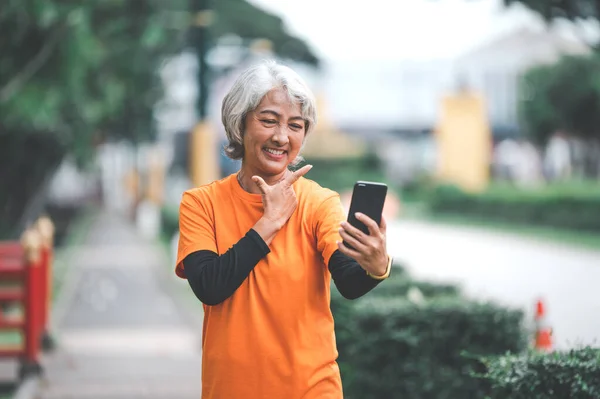 Elderly white haired Asian woman talking Video call while exercising in the park in the morning.