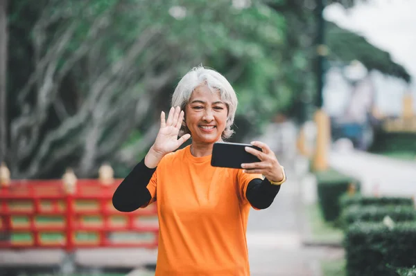 Elderly white haired Asian woman talking Video call while exercising in the park in the morning.