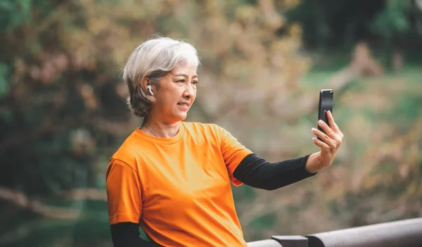 Elderly white haired Asian woman talking Video call while exercising in the park in the morning.