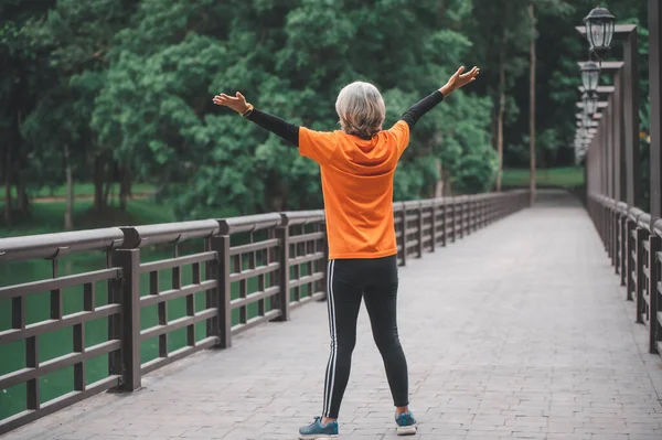 elderly white-haired Asian woman spreading her hands to breathe in the fresh air while exercising in the park early in the morning.