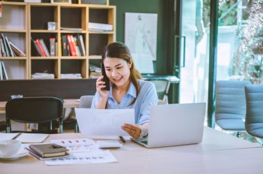 İşadamı Asyalı kadın dizüstü bilgisayarla masasında oturuyor cep telefonu kullanıyor müşterilerle sohbet ediyor ya da finansal tavsiyelerde bulunuyor..