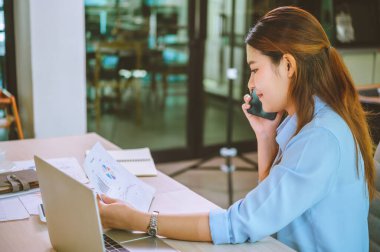 İşadamı Asyalı kadın dizüstü bilgisayarla masasında oturuyor cep telefonu kullanıyor müşterilerle sohbet ediyor ya da finansal tavsiyelerde bulunuyor..