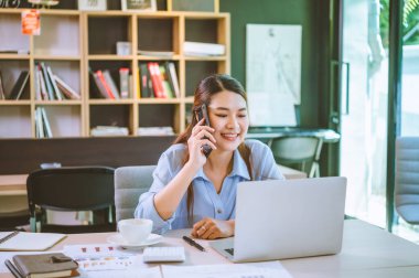 İşadamı Asyalı kadın dizüstü bilgisayarla masasında oturuyor cep telefonu kullanıyor müşterilerle sohbet ediyor ya da finansal tavsiyelerde bulunuyor..