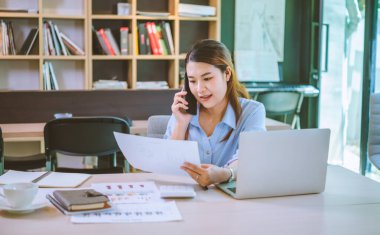 İşadamı Asyalı kadın dizüstü bilgisayarla masasında oturuyor cep telefonu kullanıyor müşterilerle sohbet ediyor ya da finansal tavsiyelerde bulunuyor..