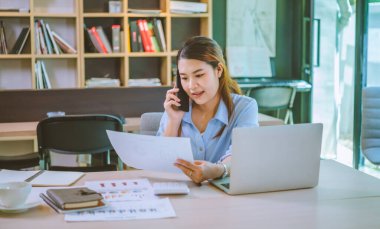 İşadamı Asyalı kadın dizüstü bilgisayarla masasında oturuyor cep telefonu kullanıyor müşterilerle sohbet ediyor ya da finansal tavsiyelerde bulunuyor..
