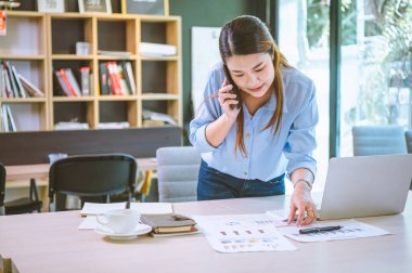 İşadamı Asyalı kadın dizüstü bilgisayarla masasında oturuyor cep telefonu kullanıyor müşterilerle sohbet ediyor ya da finansal tavsiyelerde bulunuyor..