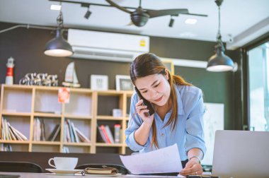 İşadamı Asyalı kadın dizüstü bilgisayarla masasında oturuyor cep telefonu kullanıyor müşterilerle sohbet ediyor ya da finansal tavsiyelerde bulunuyor..