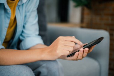 Gülümseyen Asyalı genç bir kadın cep telefonunu kullanıyor sosyal medyayı kontrol ediyor, oyun oynuyor, internetten alışveriş yapıyor, teslimat siparişi veriyor evde dizüstü bilgisayarla koltukta otururken rahatlıyor. 