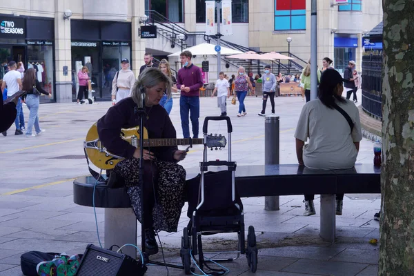 Cardiff, Wales, 18 Ağustos 2021: Genç bir bayan müzisyen sokak bankında oturuyor ve gitarını müşterilere çalıyor. St David Merkezi 'nin girişi arka planda..