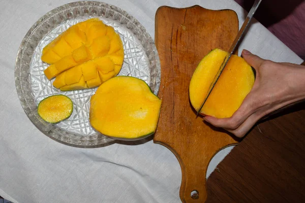 Female hands neigh ripe mango into pieces. Flatley wooden cutting board, tablecloth, knife