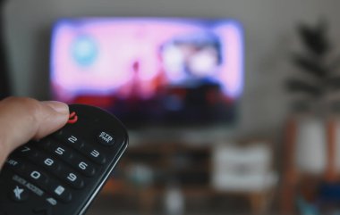 Hand holding remote control of a television. Hands pointing to tv screen set and turning it on or off select channel watching tv on his sofa at home in the living room relax.