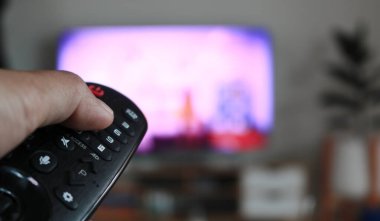 Hand holding remote control of a television. Hands pointing to tv screen set and turning it on or off select channel watching tv on his sofa at home in the living room relax.