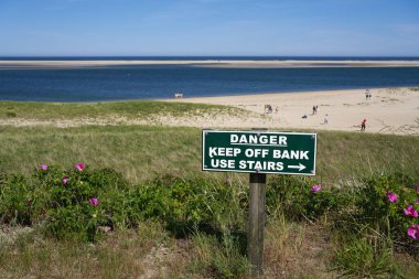 Chatham, Massachusetts 'te deniz feneri sahilinin yukarısındaki tehlike işareti. Kum tepeciğindeki parlak pembe kumsal gülleri ve diyatrandaki kumsalın keyfini çıkaran insanlar.
